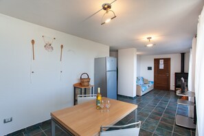 Una cocina con refrigerador, una mesa de comedor con una botella y vasos, una lámpara de pared y una puerta que da a otra habitación.