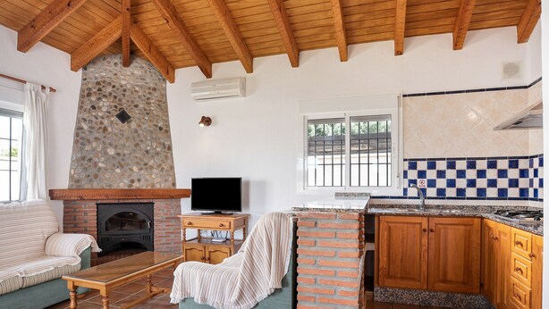 A living room with a fireplace, a television, a sofa, and a kitchen with wooden cabinets and a checkered backsplash. - Charming Casita Torre de Meca 2 with Terrace, Garden, and Wi-Fi (Los Caños de Meca)