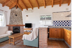 A living room with a fireplace, a television, a sofa, and a kitchen with wooden cabinets and a checkered backsplash. - Charming Casita Torre de Meca 2 with Terrace, Garden, and Wi-Fi (Los Caños de Meca)