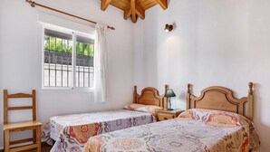 A bedroom with two beds, a wooden chair, a window with a curtain, and a nightstand with a lamp. - Charming Casita Torre de Meca 2 with Terrace, Garden, and Wi-Fi (Los Caños de Meca)