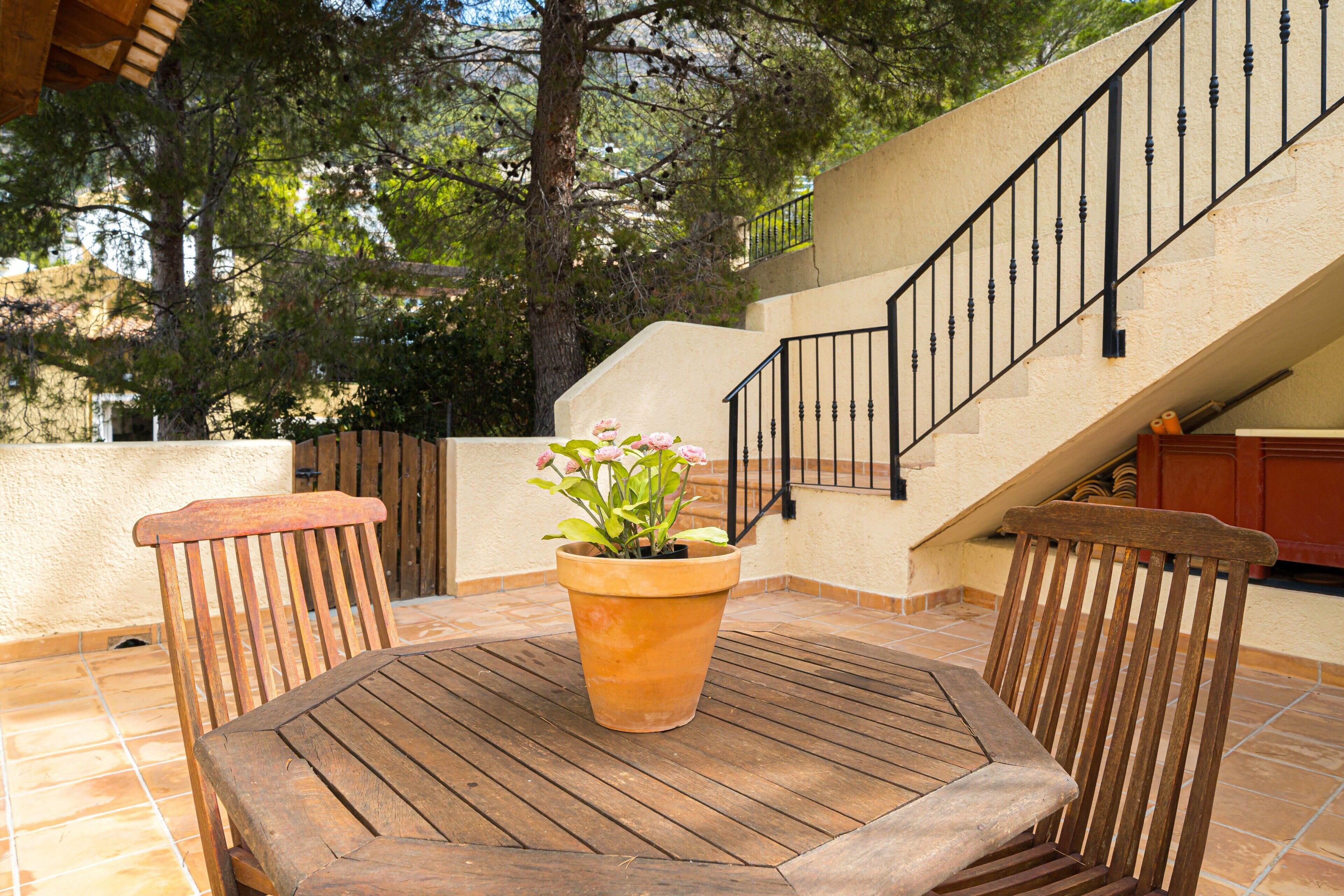 Une terrasse avec une table et des chaises en bois, un pot de fleurs et un escalier doté d’une rampe noire.