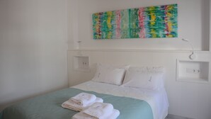 Una camera d'albergo con un letto, cuscini, una coperta verde, una testiera bianca, un'opera d'arte appesa al muro e un comodino.