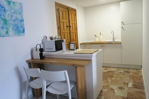 Une cuisine compacte équipée d’un four à micro-ondes, d’une bouilloire et d’un meuble en bois. On y trouve deux chaises blanches et une petite table.