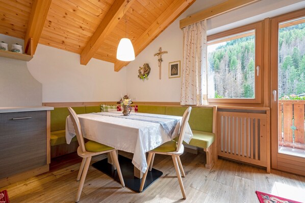 A wooden interior with a dining table, benches, a cross, and a window with a forest view. - Apartment Lüch da Mirio App Fanes with Mountain View, Garden & Wi-Fi (La Valle)