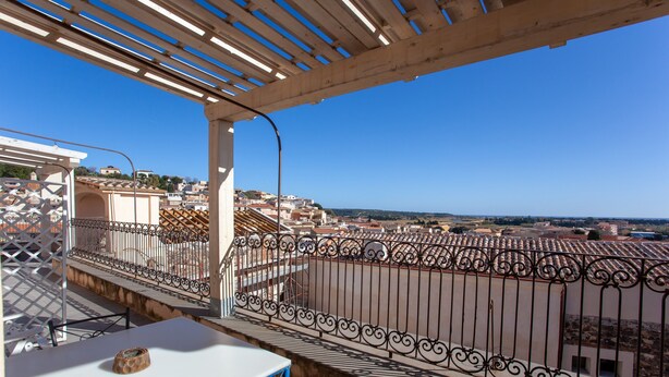 Ein Balkon mit weißem Tisch und Stühlen, einer dekorativen Schale und Blick auf eine Stadt mit Gebäuden und blauem Himmel.