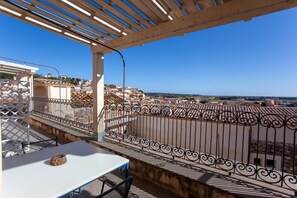 Un balcón con una mesa y sillas blancas, un cuenco decorativo y vistas a un pueblo con edificios y cielo despejado.
