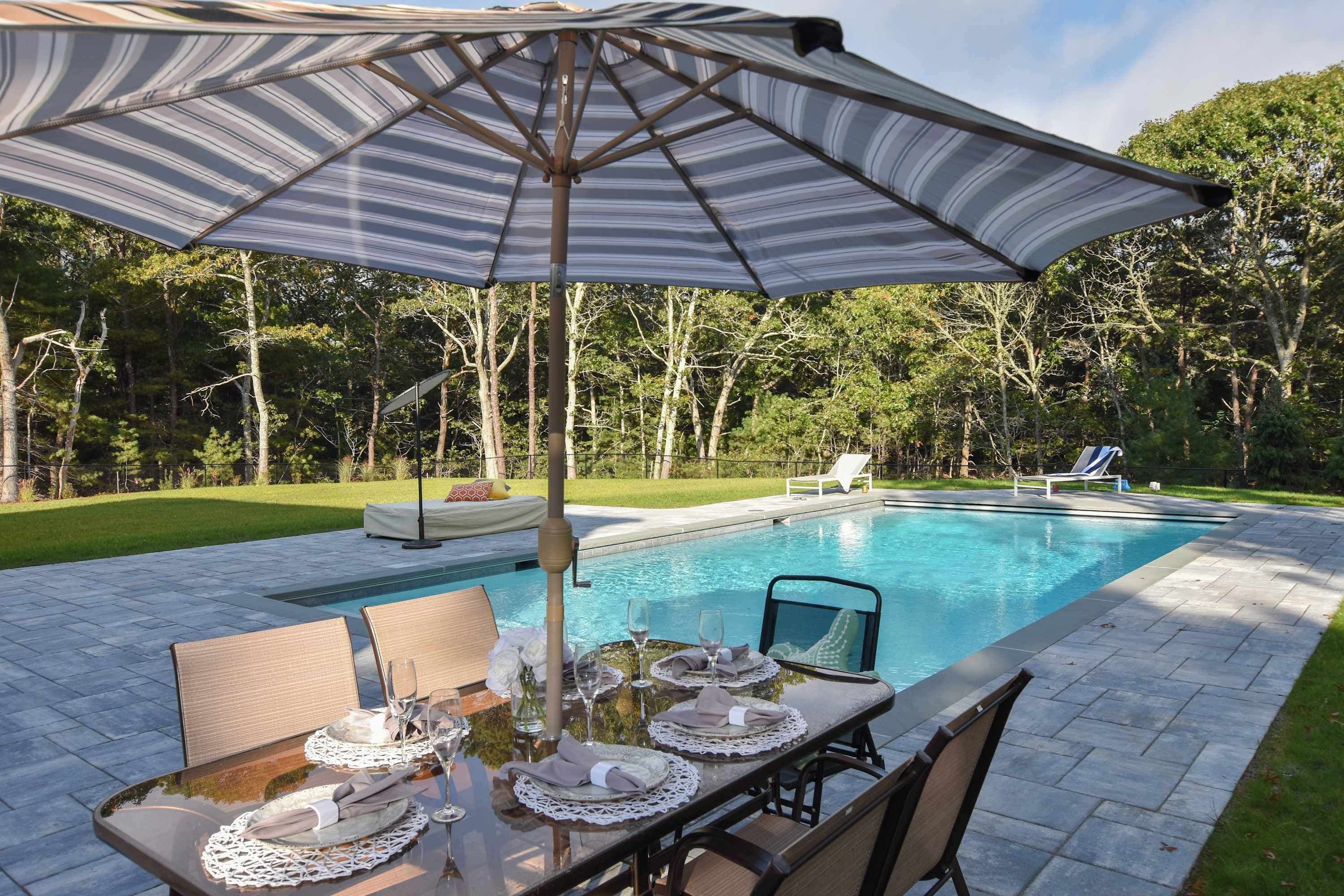 Un área de comedor junto a la piscina con una mesa preparada para dos, una sombrilla a rayas y una piscina al fondo.