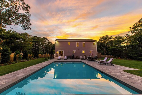 A rectangular swimming pool with a patio area, lounge chairs, and a building with a flat roof in the background.