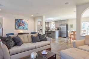 A modern living room with a sofa, coffee table, and a painting. There is a kitchen area with a refrigerator and a dining table. - Heated Saltwater Pool Hot Tub! (Eastham)