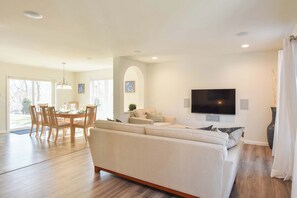 A spacious living room with a large sofa, a dining area with a wooden table and chairs, and a flat-screen TV mounted on the wall. - Heated Saltwater Pool Hot Tub! (Eastham)