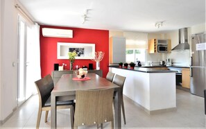 Et moderne køkken med en central ø, et spiseområde med en rød accentvæg, og en synlig aircondition.