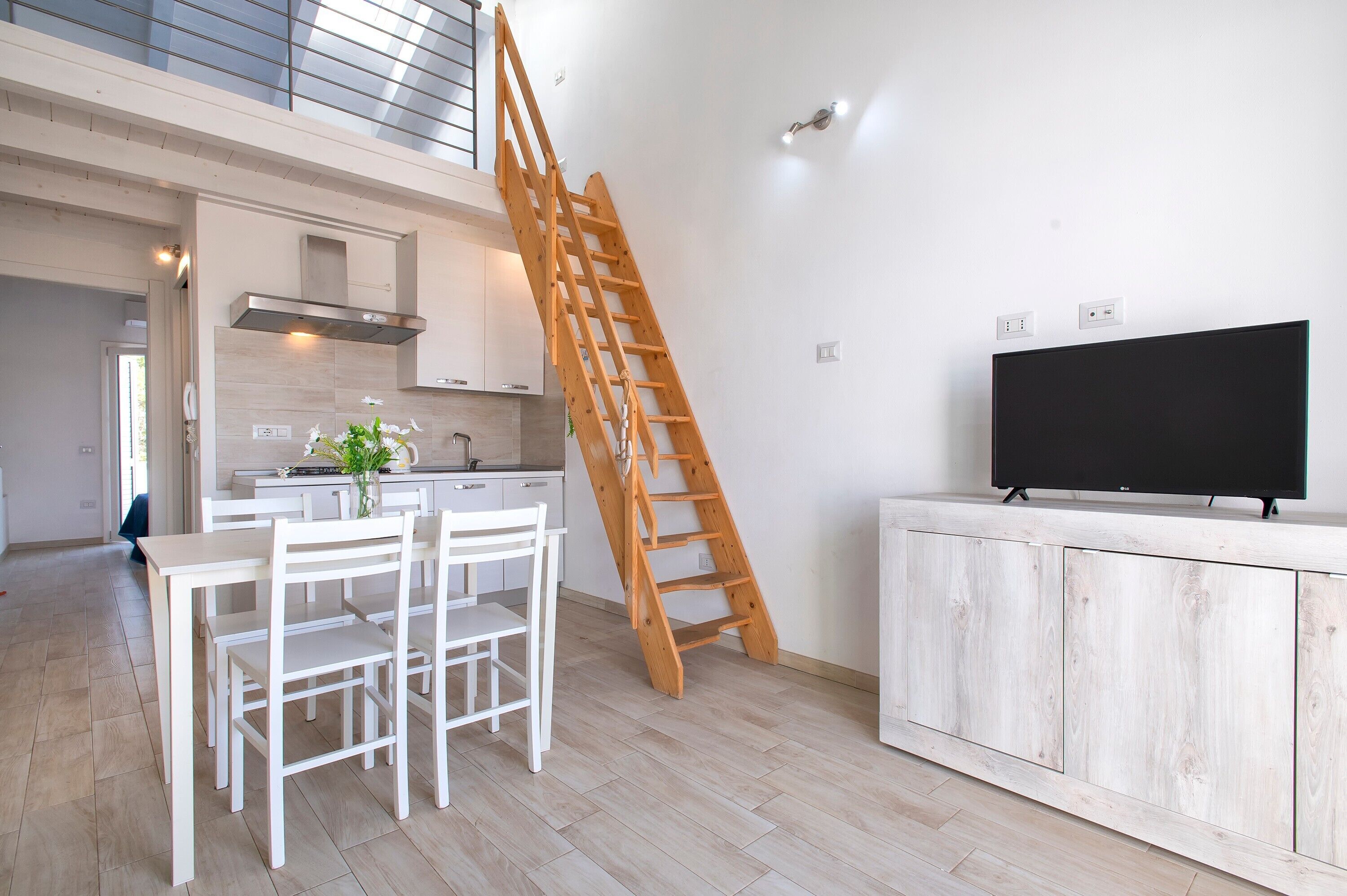 Une cuisine moderne avec une échelle en bois, un coin repas avec des meubles blancs et un téléviseur posé sur un meuble en bois.