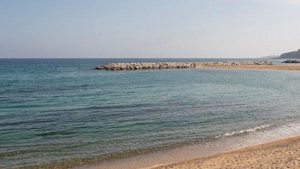 A beach with clear water, a rocky breakwater, and a distant hill. - Sunset Apartment on the Paseo de Mar in Front of the Beach with Wi-Fi (Sant Antoni de Calonge)