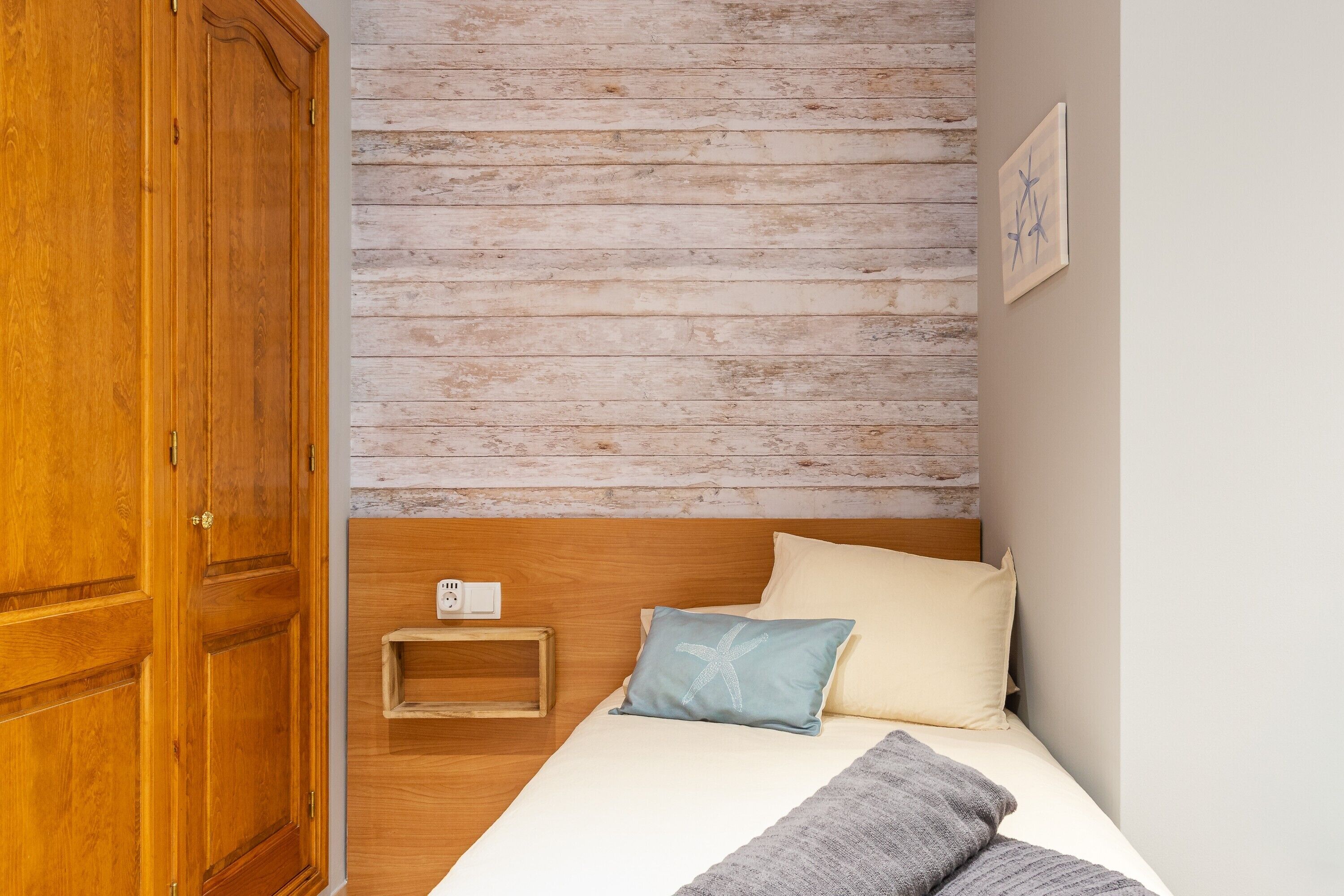 Een slaapkamer met een houten garderobe, een bed met twee kussens, een nachtkastje en een lichtschakelaar aan de muur.