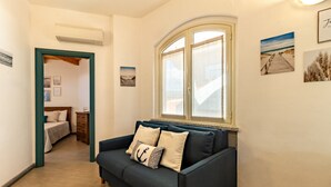 A hallway with a dark blue sofa, a wooden floor, a bed in the background, and a window with a view of the beach.