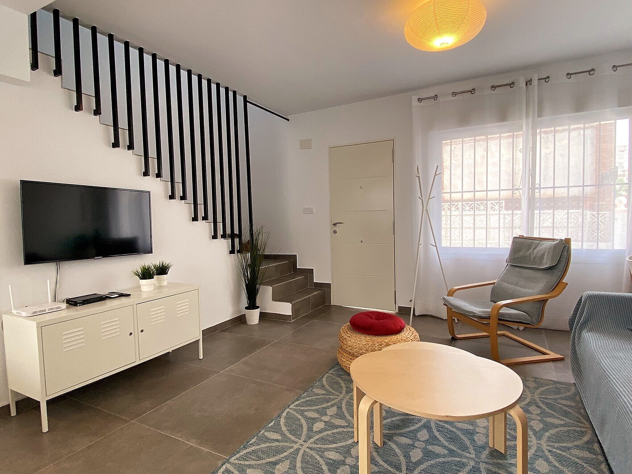 Een moderne woonkamer met een flatscreen televisie, een wit kastje, een trap met zwarte leuningen, een houten stoel, een rode kussen en een kamerbreed tapijt met patroon.