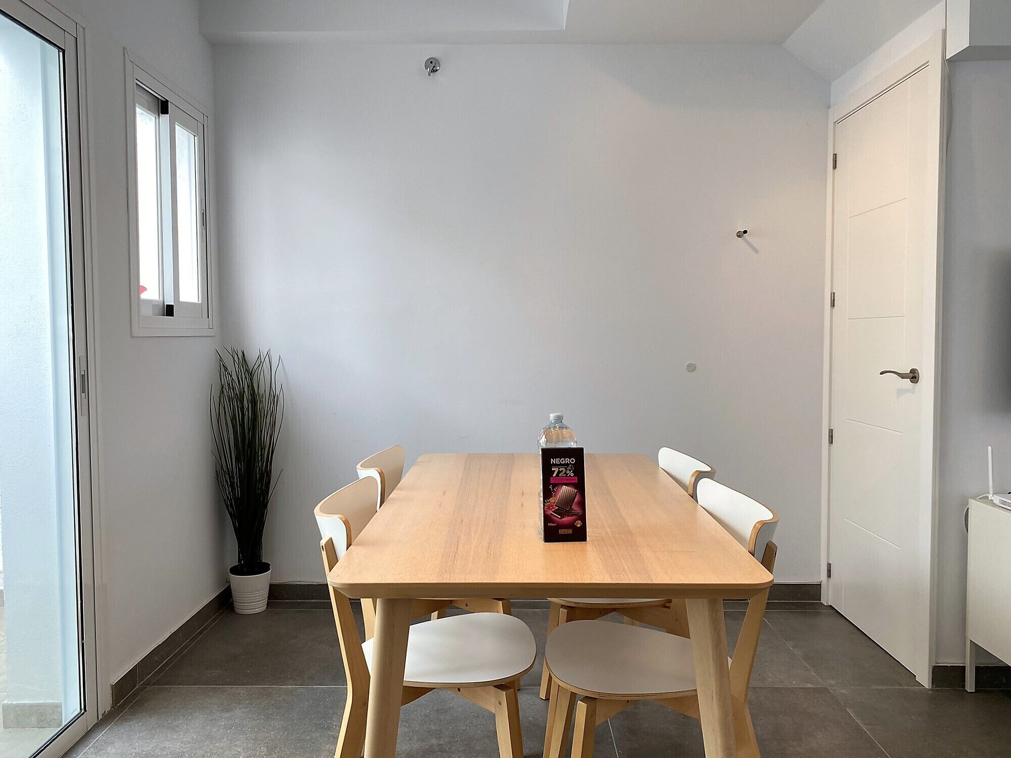 Un coin repas avec une table en bois et des chaises blanches, une plante en pot et une bouteille d’eau posée sur la table.