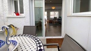 Eine kleine Terrasse mit einem gefliesten Tisch, einem Stuhl und einer kleinen Tischlampe. Eine Glastür führt zu einem Wohnzimmer mit einer Couch, einem Couchtisch und einem Sessel.