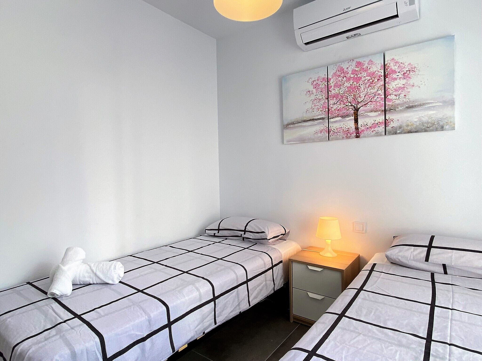 A bedroom with two beds, a nightstand with a lamp, a painting of a tree, and an air conditioner.