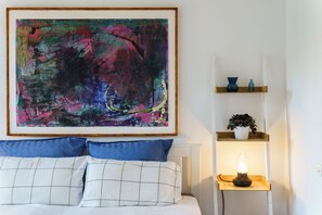 Una camera da letto con un grande quadro astratto, un letto con biancheria azzurra e bianca, un comodino con una lampada e una mensola con oggetti decorativi.