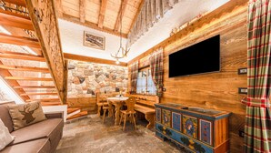 Un intérieur en bois avec un mur en pierre, un escalier en bois, un téléviseur fixé au mur, une table avec des chaises et un meuble coloré.