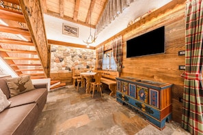 A wooden interior with a stone wall, a wooden staircase, a TV mounted on the wall, a table with chairs, and a colorful cabinet.