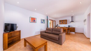A living room with a sofa, a TV, and a wooden table. There is a kitchen in the background with cabinets and a window.