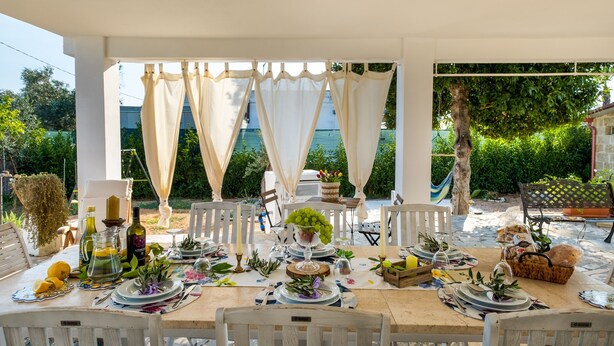 Un espace de restauration en plein air avec une table dressée pour un repas, entouré de chaises blanches, et une vue sur la verdure et un bâtiment en arrière-plan.
