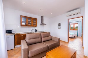 Un espacio habitable pequeño con kitchenette, un sofá, una mesa de madera y vistas a través de una puerta abierta.
