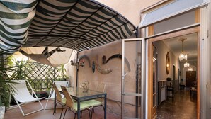 Un patio al aire libre con una mesa y sillas de vidrio, un toldo a rayas y una puerta que da a un espacio interior.