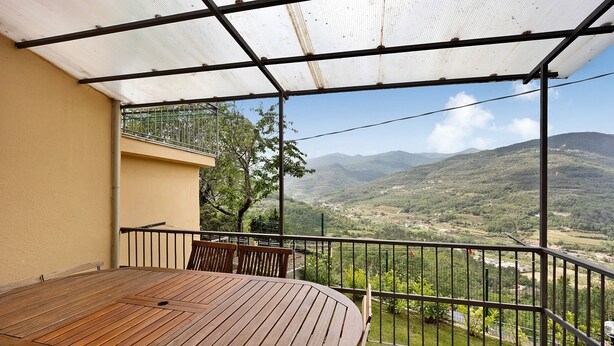 Un espacio al aire libre techado con una mesa y sillas de madera, baranda metálica y vista a un paisaje montañoso.