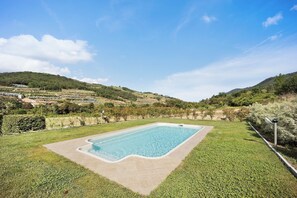 A rectangular swimming pool with a tiled edge, surrounded by a well-maintained lawn, set against a backdrop of rolling hills and a clear sky.