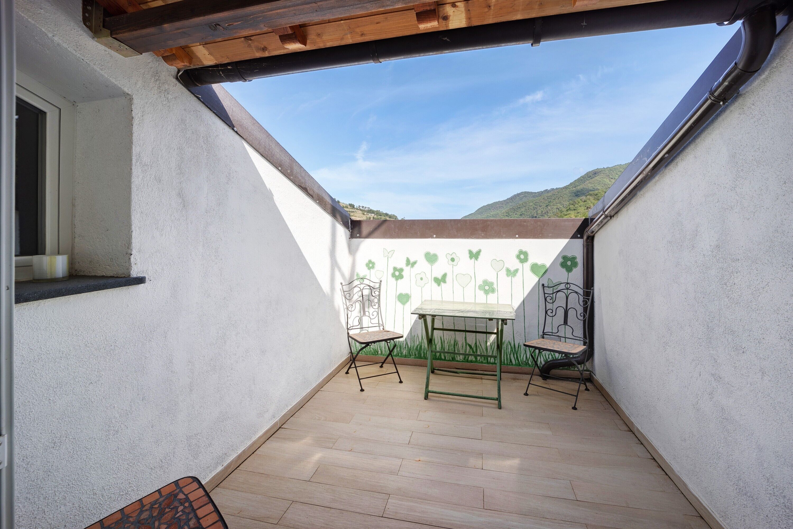 A small patio with a table and two chairs, a window, and a view of a green hill.