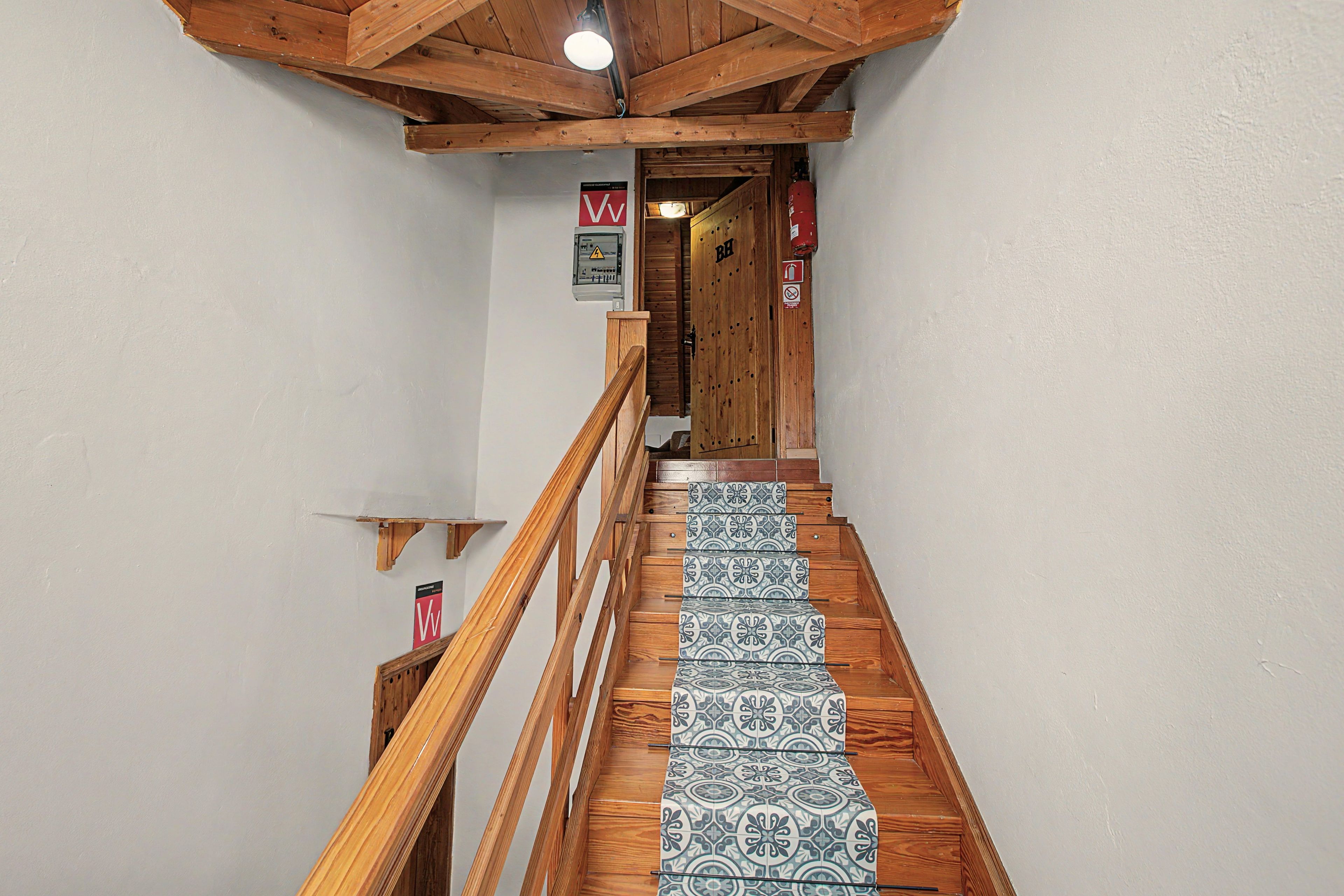 A wooden staircase with patterned tiles leading to a room with a wooden ceiling and a fire extinguisher on the wall.