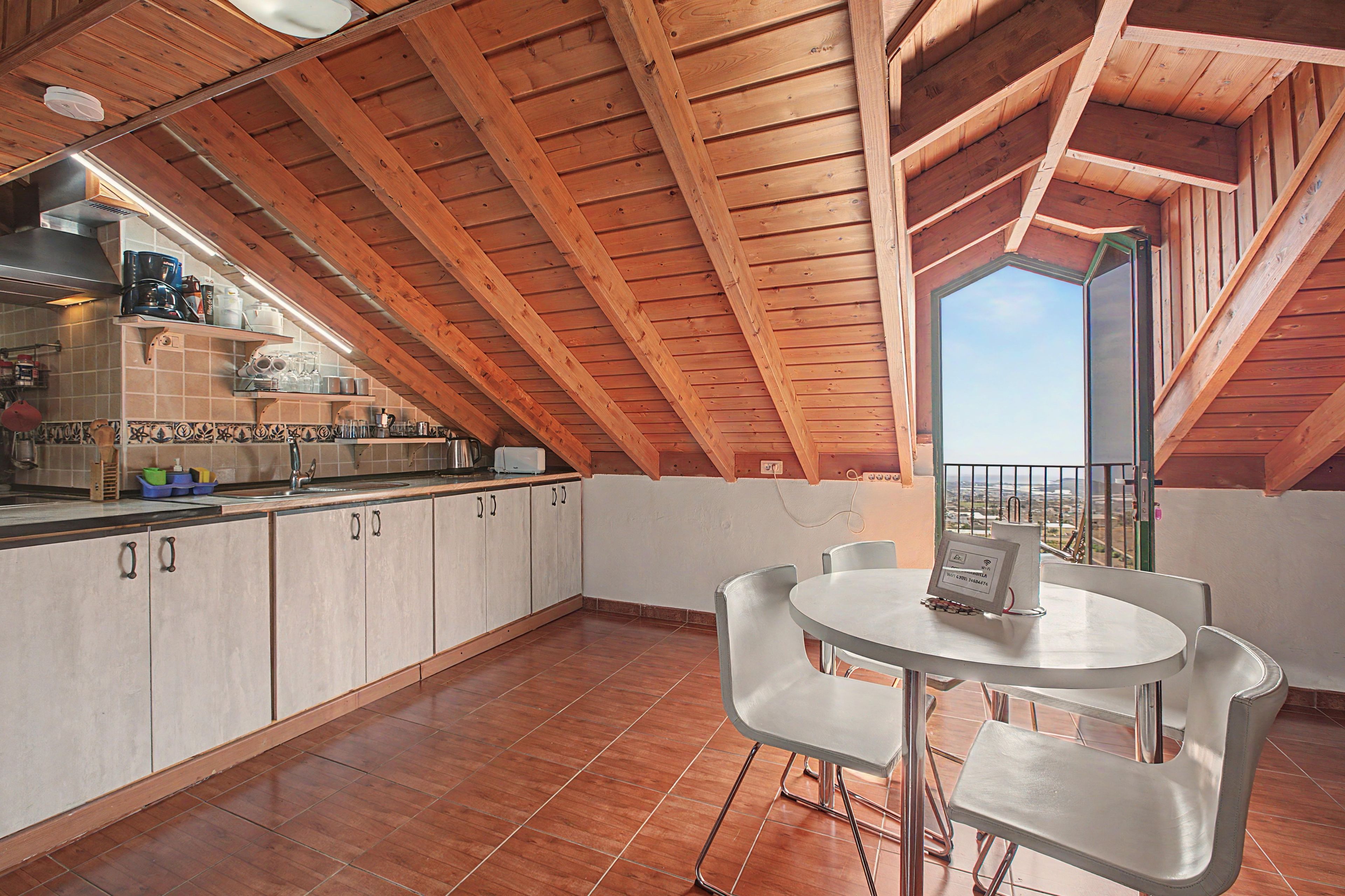 A kitchen with wooden beams, a dining area with a table and chairs, and a view through a window.