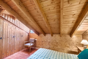 A wooden cabin room with a sloped ceiling, a bed, a chair, and a window.