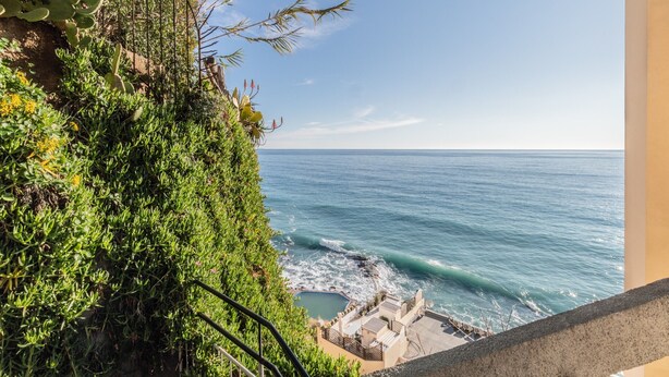 A coastal view with a rocky cliff, greenery, and a pool area.