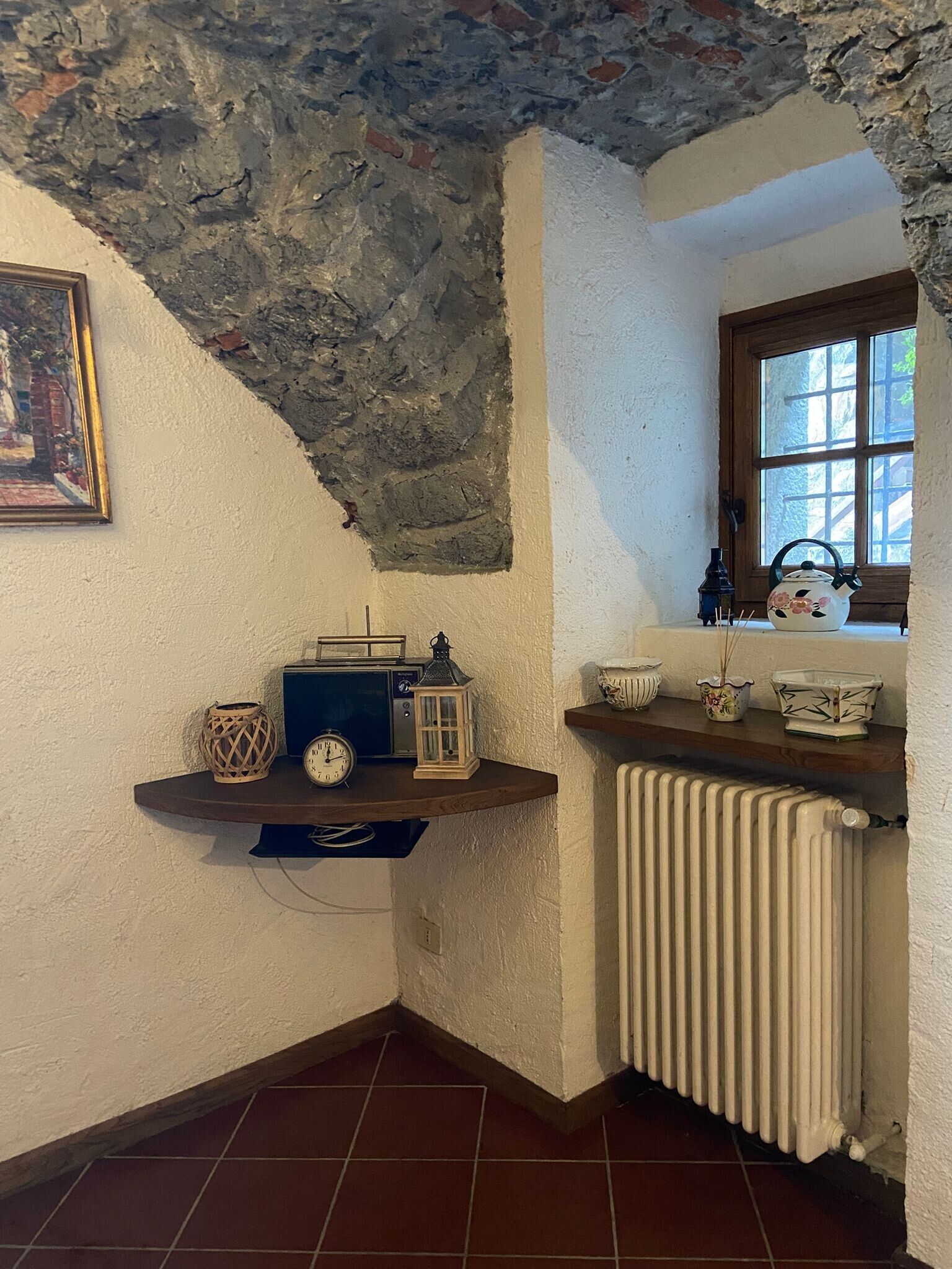 A corner room with a rustic ceiling, a wooden shelf with a radio, lanterns, and decorative items, a window with a view, and a radiator.