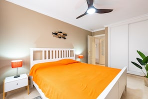 A bedroom with a white bed, orange bedding, a ceiling fan, a nightstand with a lamp, and a wall decoration.