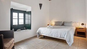 Een slaapkamer met een bed, een raam, een houten bankje, een nachtkastje en een wandlamp.