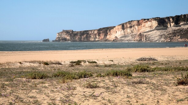 A beach with a cliff and a small structure in the distance. - Apartment 'Quinta Das Andorinhas - Nazaré' with Mountain View, Private Pool and Wi-Fi (Casais Baixo)