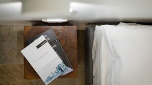 A bed with white sheets, a wooden nightstand with a book and a remote control, and a bathroom mirror.