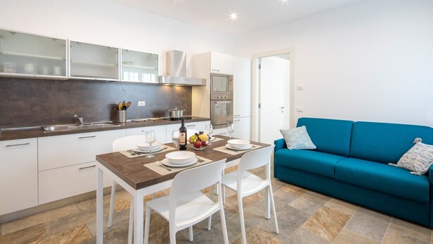 A modern kitchen with white cabinets, a dining table set for two, and a blue sofa in the living area.