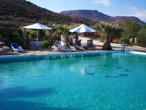A pool with clear blue water, surrounded by lounge chairs and umbrellas, with mountains in the background.