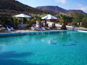Una piscina cristalina con sillas de descanso y sombrillas, rodeada de palmeras y montañas.