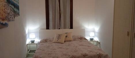 A bedroom with a bed, two bedside lamps, a window with curtains, and a wall-mounted painting.