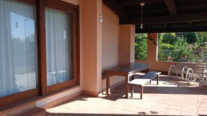 Un patio couvert avec une table et des bancs, de grandes fenêtres et une vue sur la verdure.