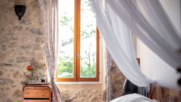 A bedroom with a stone wall, a wooden nightstand with a vase of flowers, a window with curtains, and a bed with a canopy. - "Olivier" Room – Quality B&B accommodation comes in all sizes (Fayence)