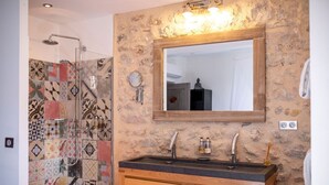 A bathroom with a stone wall, a wooden vanity, a mirror, and a shower with a tiled wall. - "Olivier" Room – Quality B&B accommodation comes in all sizes (Fayence)