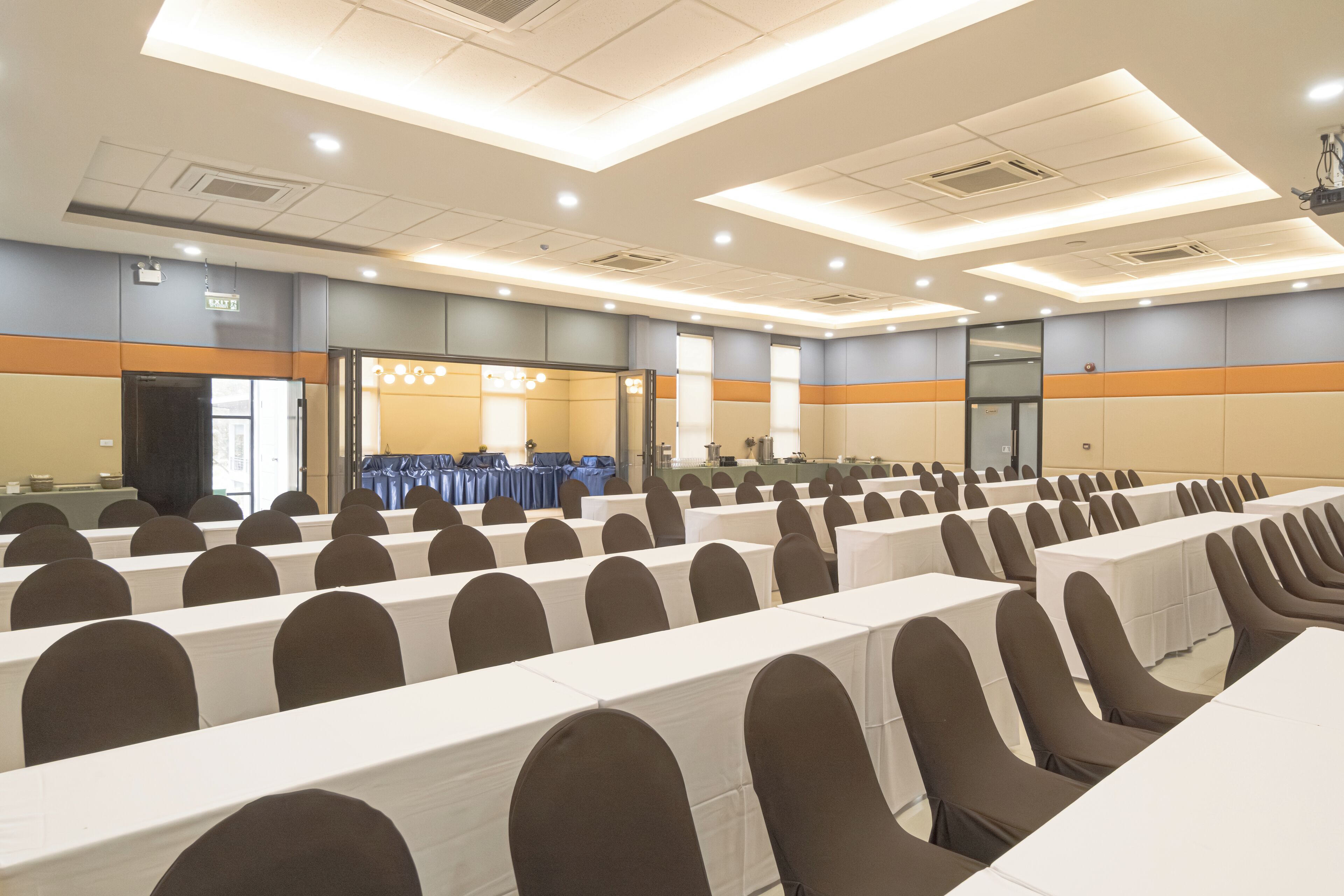 Un salón de conferencias con filas de mesas y sillas, un escenario y una puerta marcada con la leyenda 'EXIT'.